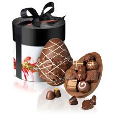 A round white gift box with a black lid and black ribbon sits behind a large chocolate egg. The egg is decorated with white lines and is open, revealing assorted chocolates inside and a few scattered outside.