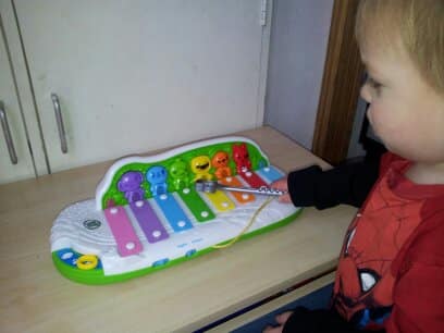 A young child in a red Spider-Man shirt plays a colourful toy xylophone with six coloured bars and smiling characters, using a yellow mallet, while sitting at a wooden table. The toy is on a light-coloured surface by a cupboard.