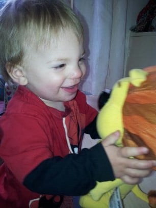 A young child with fair hair and a red shirt smiles whilst holding a yellow and orange cuddly lion toy, gazing at it happily. The background is softly lit with light curtains and furniture partially visible.