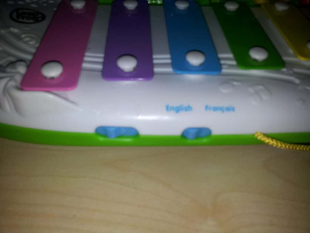 A close-up of a colourful toy xylophone with pink, purple, blue, and green bars. The words "English" and "Français" are printed in blue on the white plastic base above two blue switches. The toy rests on a light wooden surface.