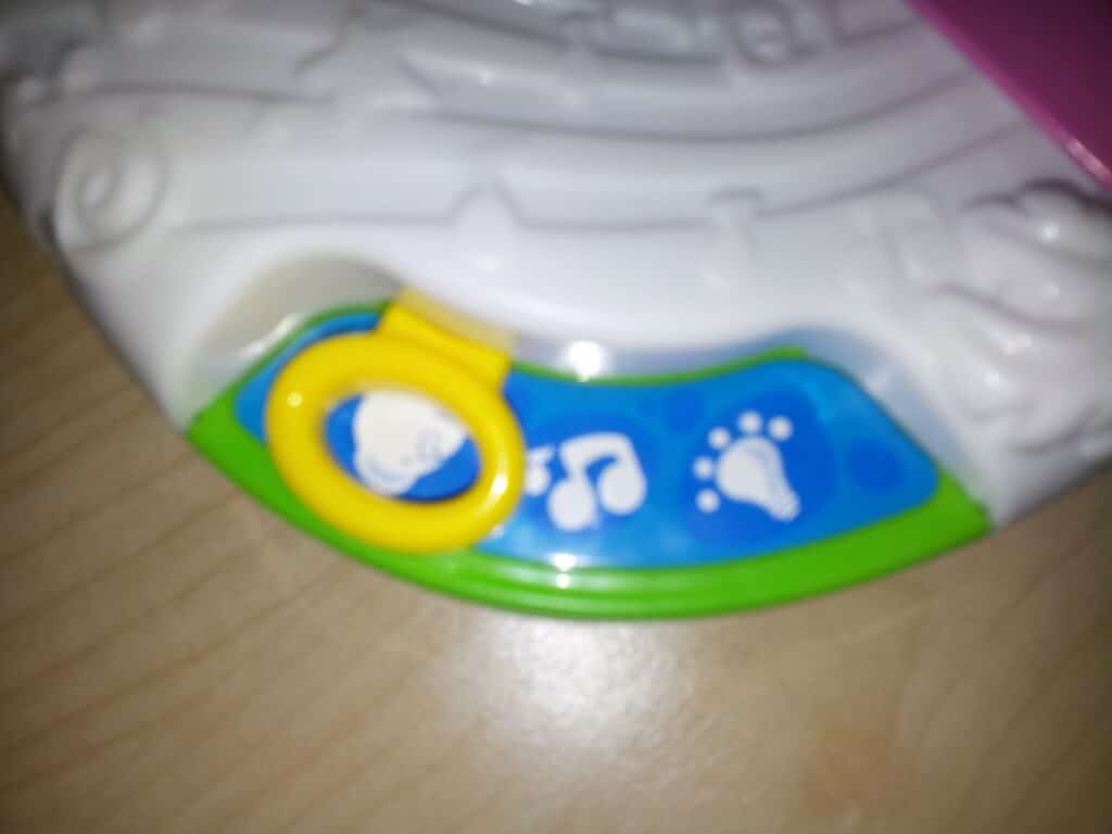 Close-up of a white plastic toy featuring a green and blue section with three icons: a bear’s face, a musical note, and a paw print. A yellow ring surrounds the bear icon. The toy rests on a light wood surface. The image is slightly out of focus.