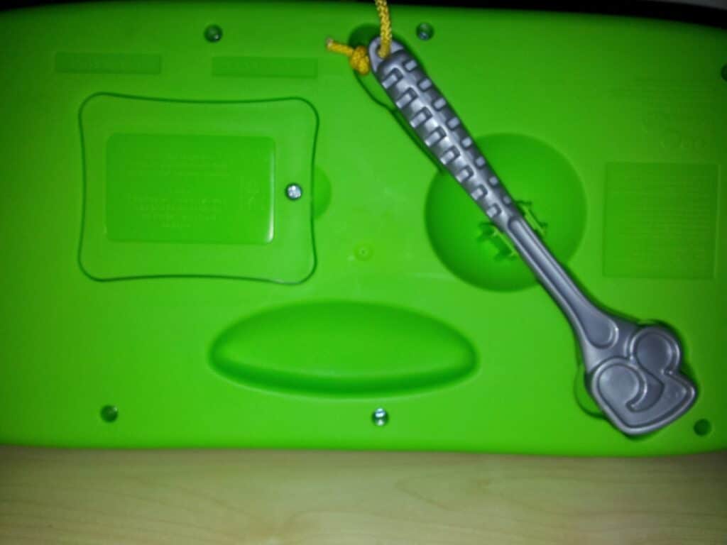 A bright green plastic object with screws and indented shapes sits on a light wood surface. A grey toy spanner with moulded details hangs from a yellow cord, attached to the object’s upper centre. The surface features several raised and recessed areas.