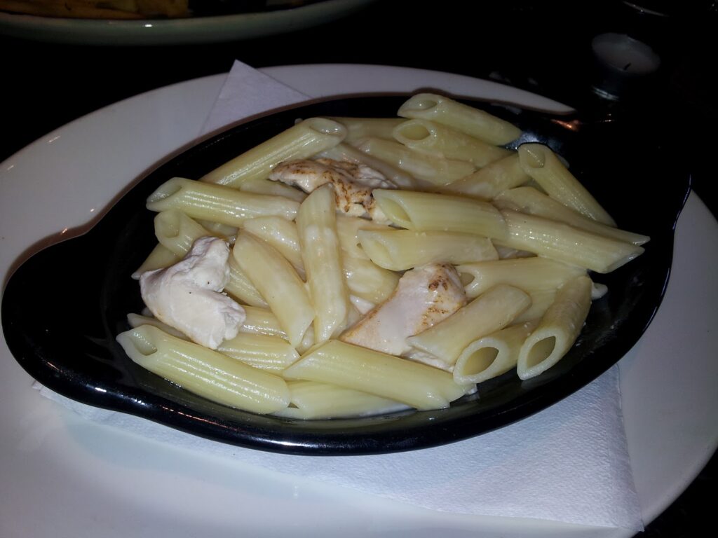 A black oval dish filled with penne pasta and chunks of chicken in a creamy white sauce. The dish sits on a white serviette on a white plate, with part of another plate visible in the background.