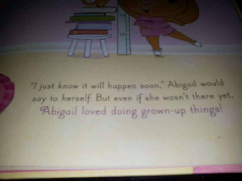 A close-up photo of a book page with the text: “‘I just know it will happen soon,’ Abigail would say to herself. But even if she wasn’t there yet, Abigail loved doing grown-up things!” The page background is yellow with a small illustration of shelves and a character’s legs.