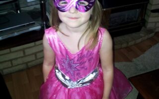 A young child stands indoors wearing a pink sparkly dress and a shiny purple and silver mask. She has light brown hair and is smiling slightly. The room has a brick fireplace, a TV on a glass stand, and framed photos on the mantelpiece.