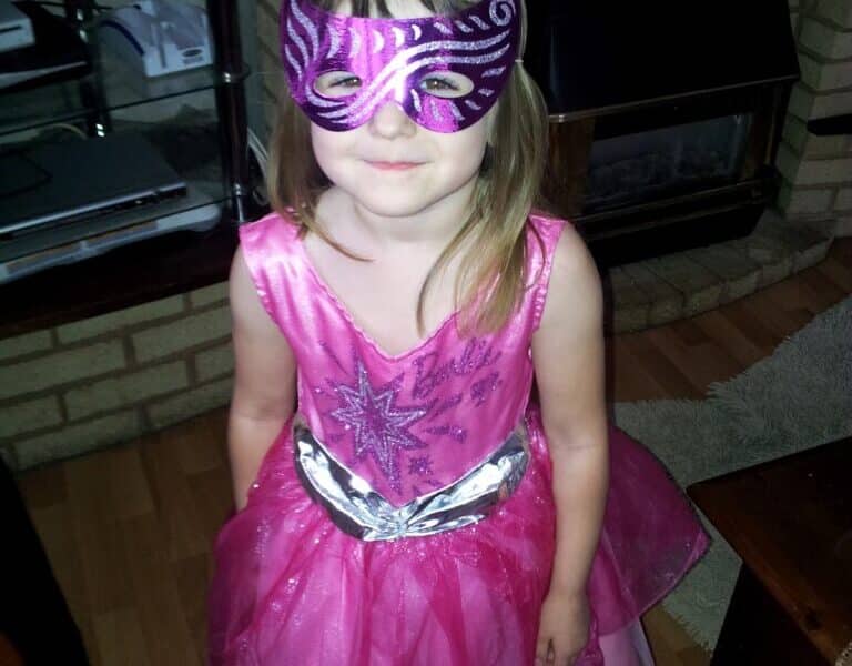 A young child stands indoors wearing a pink sparkly dress and a shiny purple and silver mask. She has light brown hair and is smiling slightly. The room has a brick fireplace, a TV on a glass stand, and framed photos on the mantelpiece.