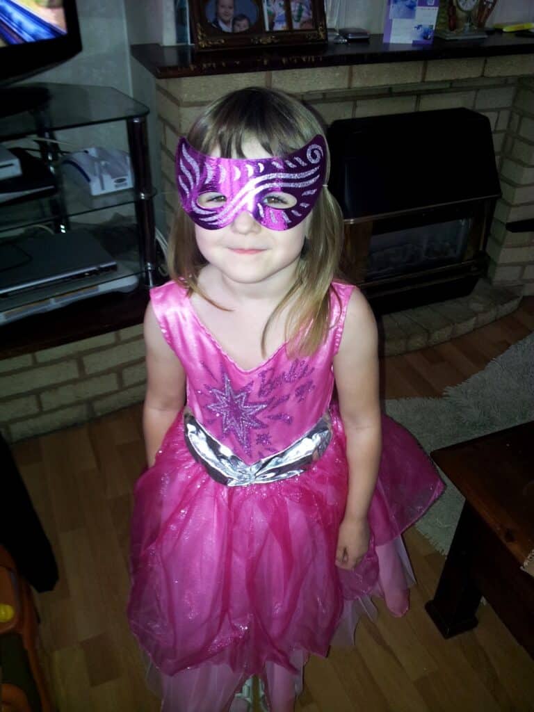 A young girl stands indoors wearing a sparkly pink dress with a silver star and a matching pink and silver mask. She has light brown hair with a fringe. The room has a fireplace, a glass shelf with items, and wooden flooring.