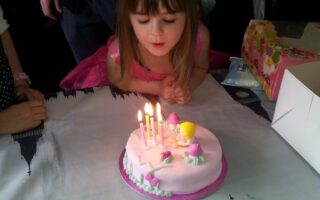 A young girl in a pink dress leans over a round, pink birthday cake with five lit candles, preparing to blow them out. The cake is decorated with pastel figures. She is indoors, at a table, with a present and adults nearby.