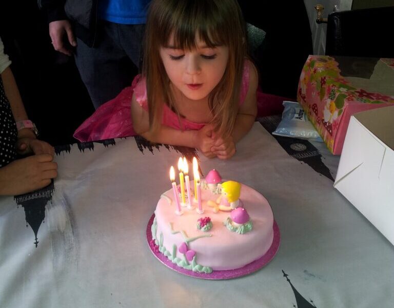 A young girl in a pink dress leans over a round, pink birthday cake with five lit candles, preparing to blow them out. The cake is decorated with pastel figures. She is indoors, at a table, with a present and adults nearby.