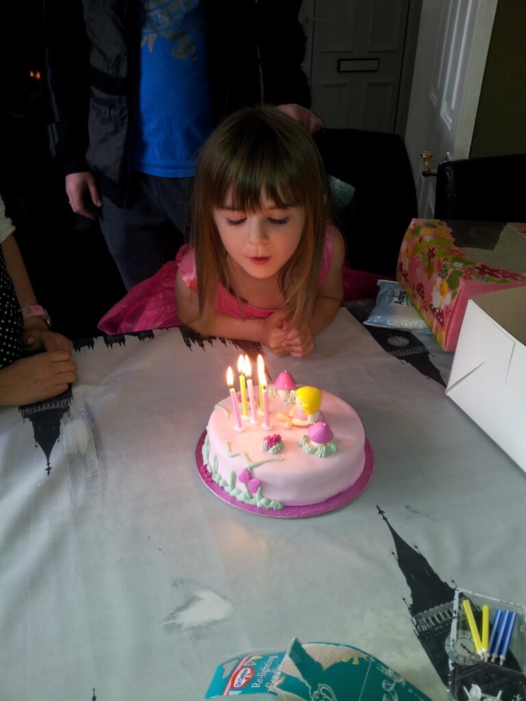 A young girl with brown hair and a pink dress leans forward to blow out five lit candles on a round, pink birthday cake with small decorations. She is surrounded by adults and presents, and the cake is placed on a table with a white tablecloth.