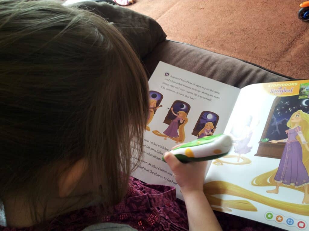 A young child sits on a sofa reading a "Tangled" storybook. The child holds a green and white reading pen over the book, which features colourful illustrations of the Rapunzel character from the Disney film. Brown carpet and a cushion are visible in the background.