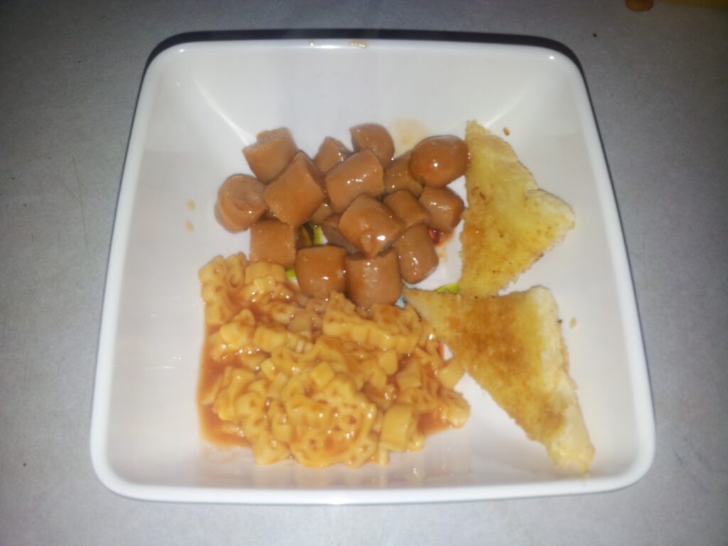 A square white plate with three foods: chopped cooked frankfurters, two triangular slices of toasted cheese sandwich, and a serving of macaroni in tomato sauce. The items are arranged separately on the plate, which sits on a light surface.