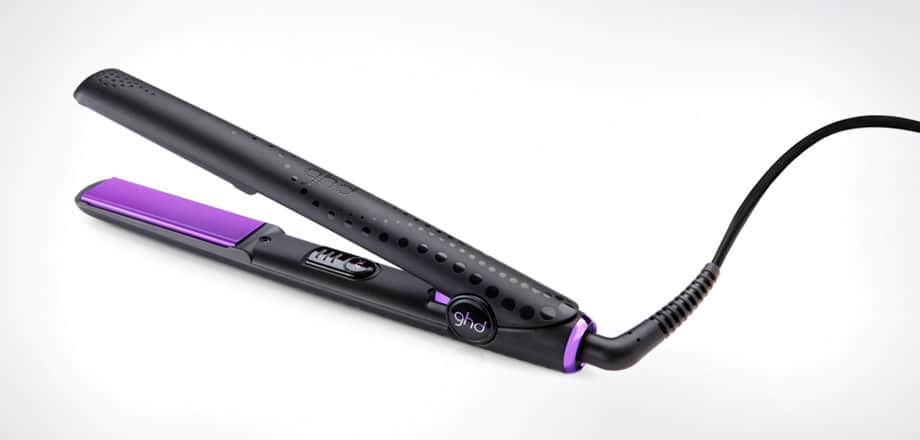 A black hair straightener with purple ceramic plates lies open on a white surface. It has a power button and an indicator light on the inner handle, with a thick black power lead attached to the end. The brand logo “ghd” is visible near the hinge.