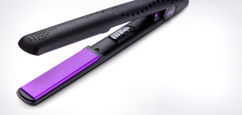 A black hair straightener with purple ceramic plates is shown open on a white background. The brand “ghd” is visible on the handle. The device has a power switch and vented holes near the tip for heat dissipation.