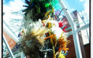 A colourful, intricate sculpture made from various objects such as straws, brushes, cable ties, and plastic items hangs indoors near a glass ceiling. Sunlight streams in, and a red-brick building is visible outside through the glass panels.