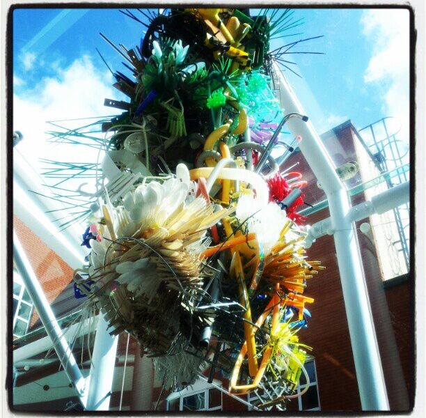 A colourful, intricate sculpture made from various objects such as straws, brushes, cable ties, and plastic items hangs indoors near a glass ceiling. Sunlight streams in, and a red-brick building is visible outside through the glass panels.