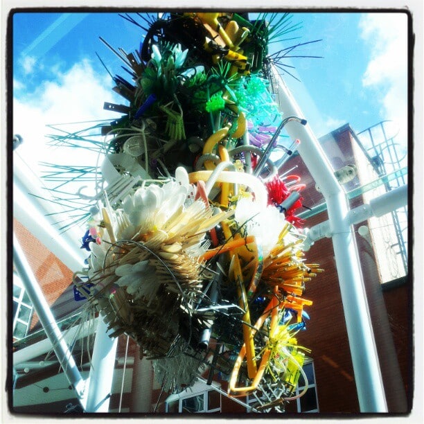 A colourful, intricate sculpture made from various objects such as straws, brushes, cable ties, and plastic items hangs indoors near a glass ceiling. Sunlight streams in, and a red-brick building is visible outside through the glass panels.