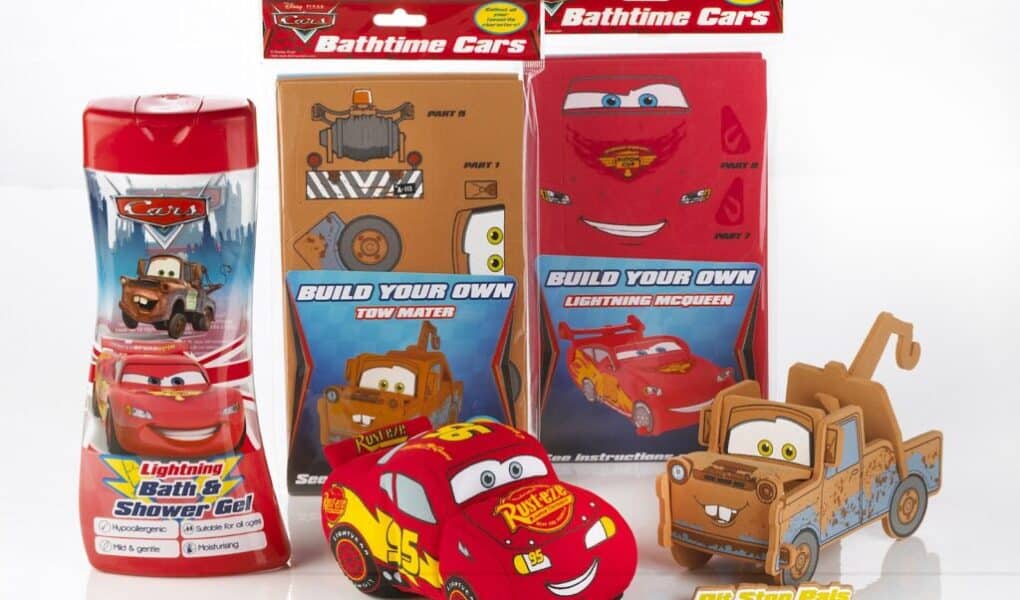 A display of Disney Cars themed bath and play products: a red bottle of Lightning Bath & Shower Gel, two flat-packed "Build Your Own" cardboard kits for Mater and Lightning McQueen, a 3D Lightning McQueen toy, and a 3D Mater toy, all on a white background.