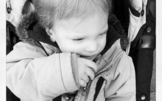 A black-and-white photo of a young child sitting in a pushchair, wearing a hooded jacket with a furry trim. The child looks down and to the side, with one hand touching their mouth and a gentle, thoughtful expression on their face.