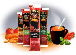 Four Heinz Squeeze & Stir soup sachets stand amid fresh vegetables, with a steaming black mug of tomato soup and a spoon beside them. The sachets include Cream of Tomato and Tomato with Basil flavours. Tomatoes, carrots, and herbs decorate the scene.