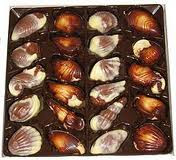 A box of assorted Belgian chocolates shaped like seashells and sea creatures. The chocolates appear marbled in brown and white, neatly arranged in a dark plastic tray with individual compartments.