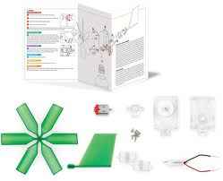 A do-it-yourself wind turbine kit is shown. The kit includes green plastic blades, a small motor, white plastic parts, wires, screws, a green sheet, and an illustrated instruction manual with assembly diagrams and coloured text.
