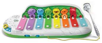 A colourful children’s xylophone toy with seven keys in rainbow colours, each with a smiling cartoon character above it. A toy microphone and a speaker are attached on the right side. The toy base is white with green detailing.