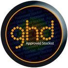 A round black badge with “ghd” written in lowercase gold dots at the centre. Below "ghd" are the words "Approved Stockist" in smaller white text. The badge has a thin gold outline and a subtle light reflection on the top right edge.