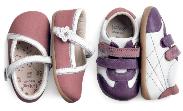 Two pairs of baby shoes are shown: one pink pair with white straps and a flower detail, and one sporty pair in white and purple with velcro fastenings and tan soles. Both pairs are soft-soled and designed for infants or toddlers.