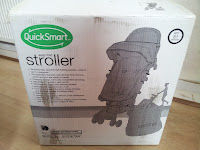 A cardboard box with "QuickSmart Stroller" text and a sketch of the stroller printed on it stands on a wooden floor. The box lists pram features and has a green logo in the top left corner. The photo is taken indoors near a radiator.