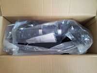 A cardboard box containing a large black electronic device wrapped in clear plastic and foam for protection. The device appears new and partially visible, with a white instruction sheet placed on top inside the packaging.