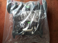A folded black pushchair is wrapped in a clear plastic bag, placed on a reddish-brown carpet. Parts of the wheels and frame are visible through the plastic, along with part of a white logo and a small green label.