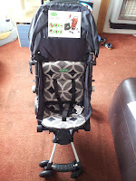 A black and grey baby pushchair with a geometric patterned seat is positioned on a reddish-brown carpet. The pushchair has a harness, a footrest, and a sign with product information attached to the canopy. A sofa and other household items are visible in the background.
