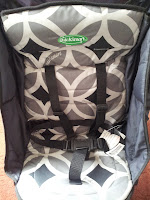 A close-up of a Graco child car seat with a geometric grey and white patterned fabric. The seat features black safety harness straps and visible buckles. The padding and sides are dark, and a white tag is attached near the lower right side.