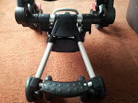 The image shows the base of a pram or pushchair on a brown carpet. The pram has two silver metal bars, black plastic joints, and small wheels, with a textured footrest near the bottom of the frame.