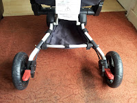 A close-up of the bottom section of a pushchair showing two large black wheels with red brakes, a grey metal frame, and a black fabric basket underneath, all situated on a reddish-brown carpeted floor near a door.