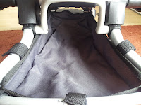 A close-up view of the storage basket underneath a pushchair. The basket is made of dark fabric and framed by light grey metal bars, with black straps attached at the sides. The inside of the basket is empty, and the floor is partially visible in the background.