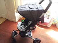 A black and grey pushchair with a geometric patterned seat is placed indoors on a brown carpet near a window. Some toys, a box, and a curtain are visible in the background, with sunlight streaming in.