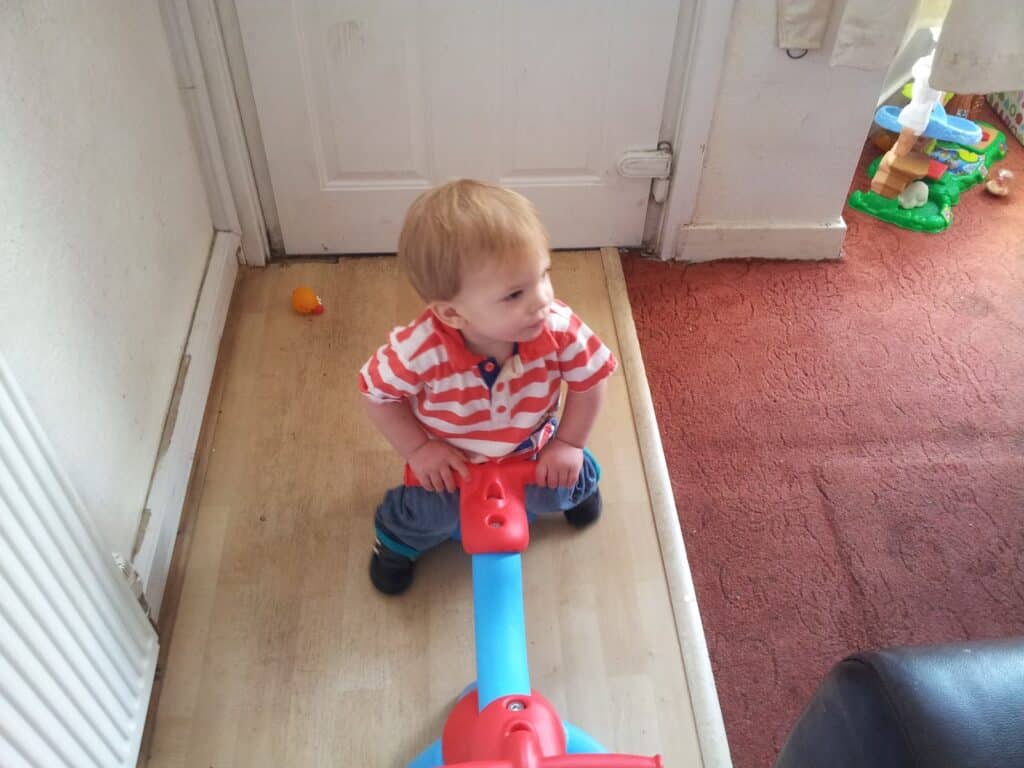 A young child with short blonde hair, wearing a red-and-white striped shirt and blue trousers, sits on a colourful plastic seesaw indoors near a white door. A toy and a play area with a green base are visible on the red carpet nearby.