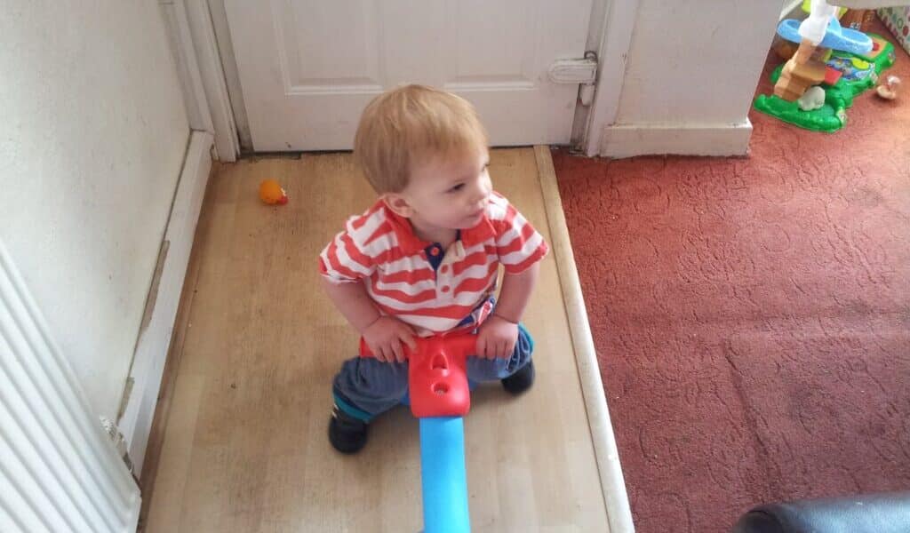 A small child with light brown hair wearing a red-and-white striped shirt and blue trousers sits on a blue and red seesaw toy, looking to the side. The child is indoors, near a white door and a red carpeted area with a toy in the background.