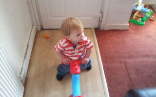 A small child with light brown hair wearing a red-and-white striped shirt and blue trousers sits on a blue and red seesaw toy, looking to the side. The child is indoors, near a white door and a red carpeted area with a toy in the background.