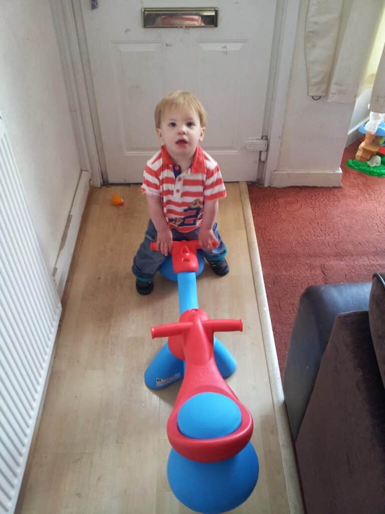 A young child with light brown hair wearing a red, white, and blue striped shirt and jeans sits on a colourful plastic seesaw indoors, positioned in a narrow hallway with a closed white door behind him. The floor is light wood, and there’s red carpet to the side.