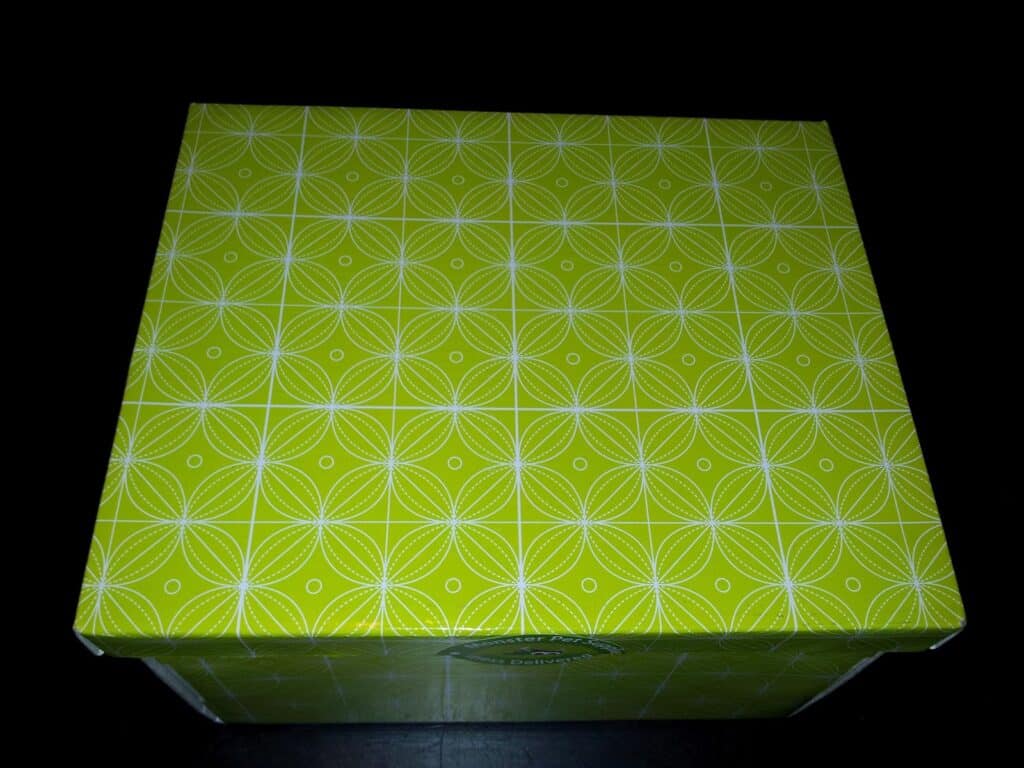 A square box with a bright green lid featuring a repeating geometric pattern of overlapping white circles and lines, creating a floral effect. The box is photographed from above against a dark background.