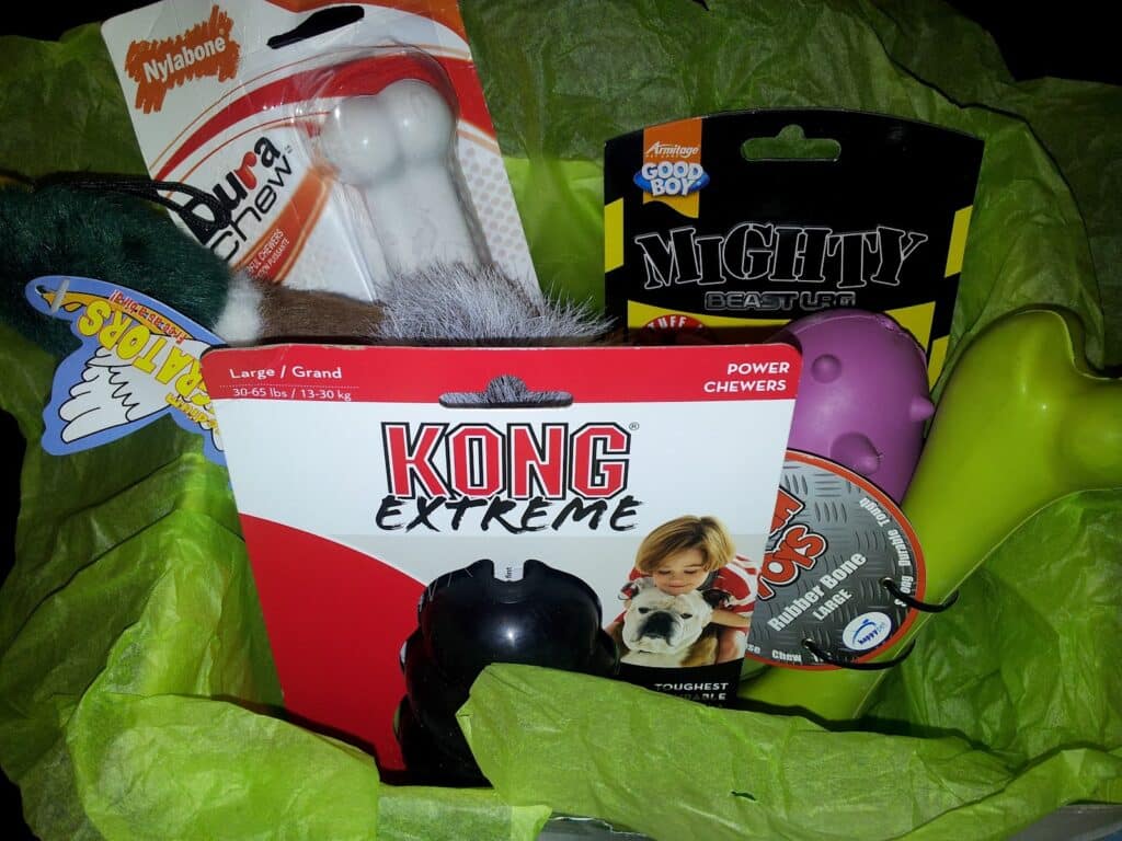 A gift hamper lined with green tissue paper contains various dog toys and treats, including a white Nylabone, a squeaky furry toy, a black KONG Extreme toy, a purple rubber toy, a green rubber bone, and a tin labelled "Antlerbone.