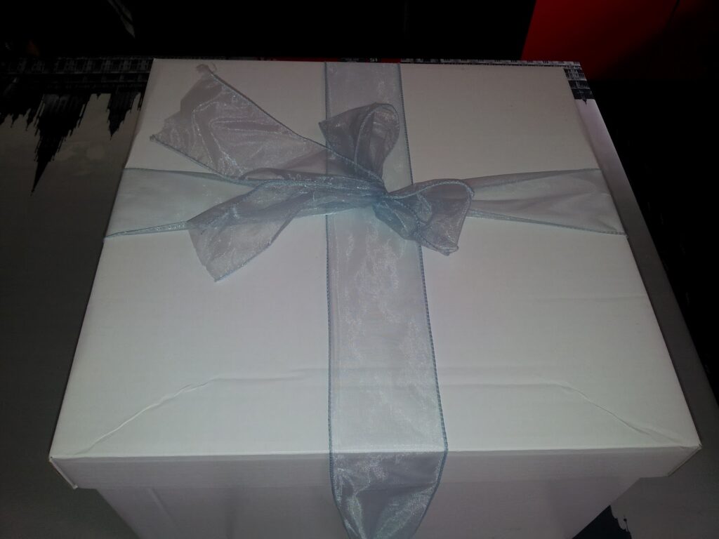 A white square gift box is topped with a sheer, light blue ribbon tied in a bow. The box sits on a reflective surface, with a dark background partially visible at the top. The overall lighting is soft, creating subtle shadows.