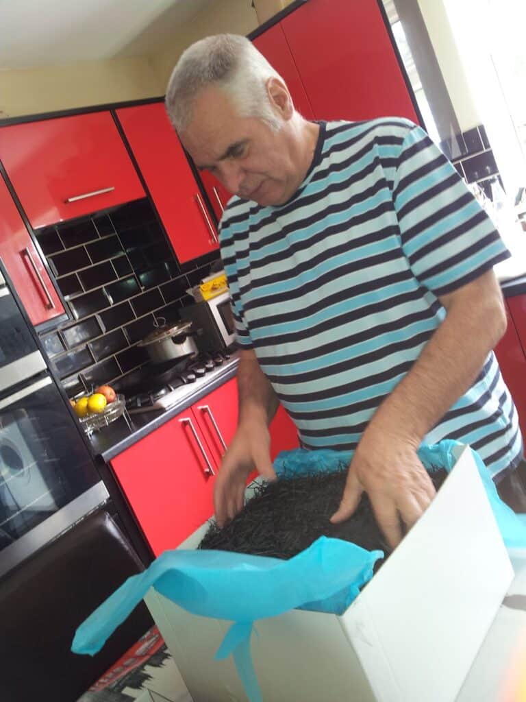 An older man in a striped shirt stands in a kitchen with red cupboards, looking down while touching black shredded paper inside a white box lined with blue tissue paper. A bowl of fruit and cooker are visible in the background.