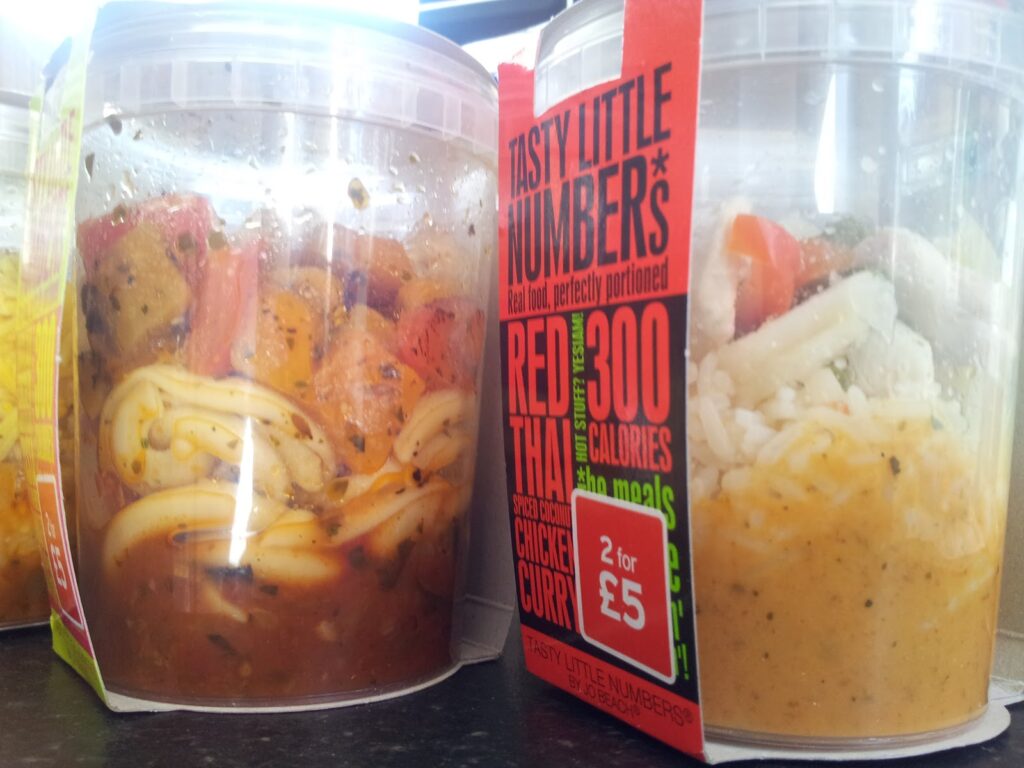 Two clear plastic tubs of ready meals are shown. The left contains noodles and vegetables in a red sauce. The right tub, labelled “Tasty Little Numbers Red Thai Curry,” displays the meal inside, plus bold calorie and price information on a red sleeve.