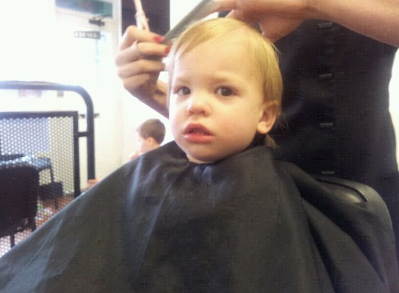 A young child with fair hair sits in a barber’s chair, covered with a black cape, looking slightly alarmed. An adult’s hands are cutting the child’s hair. The background shows another child and salon furniture out of focus.