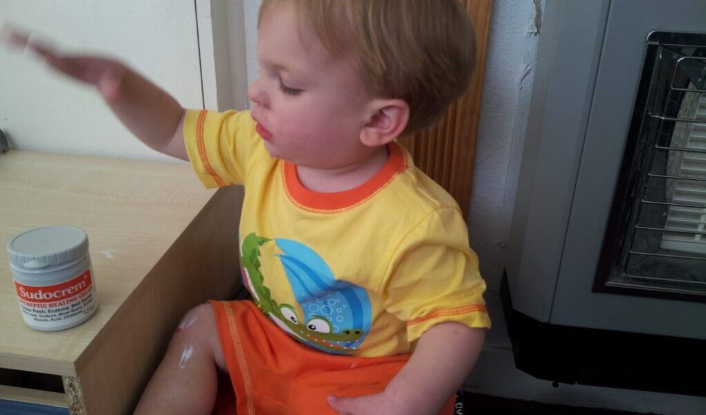 A toddler in a yellow and orange outfit sits near a wooden shelf, reaching towards an open tub of Sudocrem. Some cream is smeared on their leg. The child has fair hair and is looking at the cream, with various objects and a heater nearby.