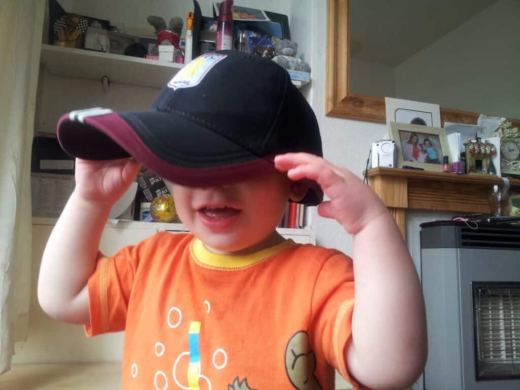 A young child wearing an orange shirt with cartoon designs smiles while pulling down the brim of an oversized black and maroon cap, partially covering their face. The background shows shelves, family photos, and various household items.